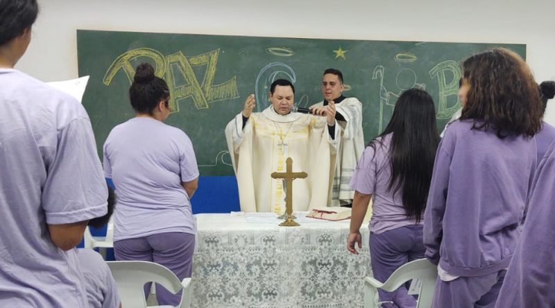 Dom Cícero, bispo auxiliar, um homem branco com vestes nos tons branco, bege e dourado, está ao centro de uma mesa com toalha branca e crucifixo, fala para as jovens com as mãos erguidas, enquanto um seminarista segura o microfone para ele. Adolescentes brancas e pardas acompanham a missa - elas utilizam blusas e calças na cor lilás.