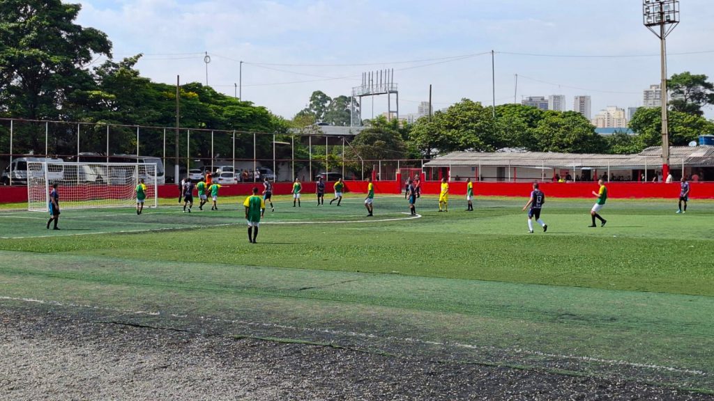 Partida de futebol em campo sintético, cercado por tela de proteção, com jogadores em ação. Arquibancadas baixas e prédios ao fundo compõem o cenário.