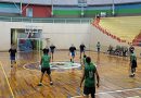 Fotografia mostra adolescentes durante partida de basquete, jogada dentro de um ginásio com quadra de madeira. O ginásio tem um formato oval, teto alto e é bem iluminado. Os jovens usam uniformes da modalidade, nas cores verde e azul.