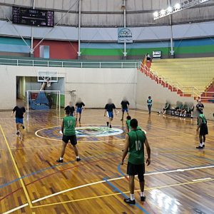 Fotografia mostra adolescentes durante partida de basquete,  jogada dentro de um ginásio com quadra de madeira. O ginásio tem um formato oval, teto alto e é bem iluminado. Os jovens usam uniformes da modalidade, nas cores verde e azul. 