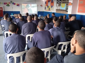 Foto mostra vários adolescentes sentados em cadeiras, numa parte do pátio do centro de atendimento, assistindo a um filme projetado no telão. As paredes da área são nas cores branca e azul e estão decoradas com bandeiras de festa junina e mensagens em cartazes sobre antirracismo