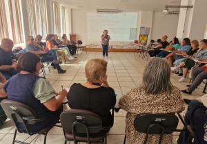 Foto mostra o auditório da DRCAP, uma sala ampla, com cadeiras distribuídas em formado de U e com os servidores sentados nelas. A parede do fundo tem a projeção de um trecho de livro e, à frente da projeção, no centro da distribuição das cadeiras, uma mulher fala ao microfone