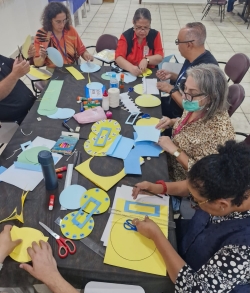 Foto mostra uma mesa em close superior, com sete adultos, entre homens e mulheres brancos e negros, sentados em cadeiras ao redor dela. Eles estão manuseando papéis coloridos, montando discos amarelos e retângulos azuis, utilizando régua e tesoura