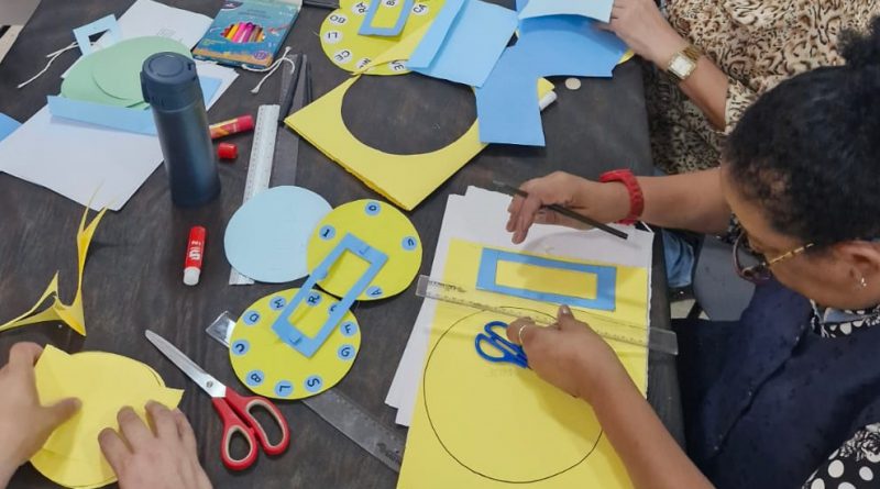 Foto mostra uma mesa em close superior, com três adultos a sua volta, que estão manuseando papéis coloridos, montando discos amarelos e retângulos azuis, utilizando régua e tesoura
