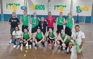 Foto mostra 8 adolescentes da equipe campeão, com uniforme nas cores verde (camiseta) e preta (calção), em formação de time, na quadra do Instituto. Junto aos jovens há cinco adultos, entre homens e mulheres. Os jovens estão com medalhas e troféu de vencedores