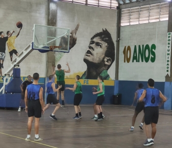 Foto mostra quadra do Instituto Neymar Jr. As paredes estão nas cores branca e azul e nelas estão pintadas uma imagem do rosto do jogador, olhando para cima; o nome do Instituto e o dizer: 10 anos. Na quadra estão dois times de basquete, com 8 jovens com uniformes nas cores verde (camiseta) e preta (calção) e azul (camisa) e preto (calção). Um dos jovens da equipe verde salta na direção da tabela de basquete