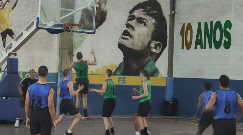 Foto mostra quadra do Instituto Neymar Jr. As paredes estão nas cores branca e azul e nelas estão pintadas uma imagem do rosto do jogador, olhando para cima; o nome do Instituto e o dizer: 10 anos. Na quadra estão dois times de basquete, com 8 jovens com uniformes nas cores verde (camiseta) e preta (calção) e azul (camisa) e preto (calção). Um dos jovens da equipe verde salta na direção da tabela de basquete