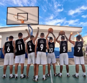 Foto mostra sete adolescentes do time de basquete em pé, de costas, utilizando uniforme nas cores branco e preto, na quadra do centro socioeducativo. Eles estão com os braços erguidos, sendo que dois deles seguram bolas d basquete. O ângulo mostra uma tabela de basquete acima deles, ainda tendo ao fundo o telhado de uma edificação. O céu está azul, ensolarado e com poucas nuvens
