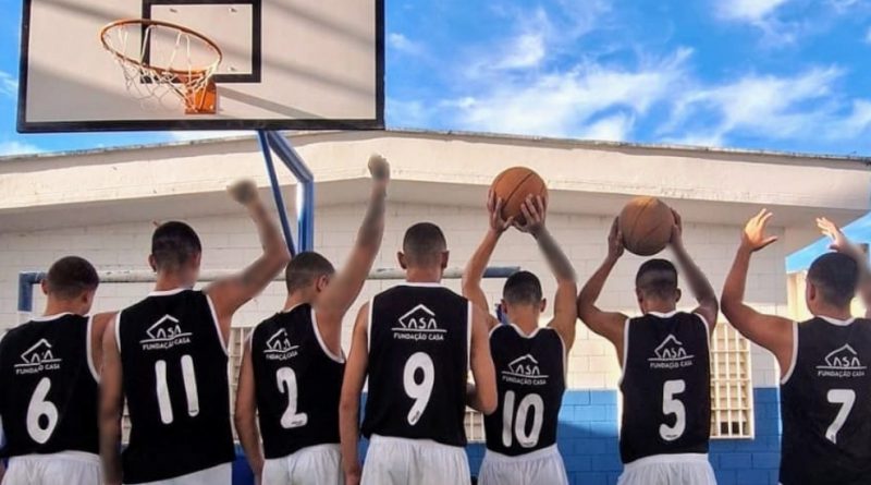 Foto mostra sete adolescentes do time de basquete em pé, de costas, utilizando uniforme nas cores branco e preto, na quadra do centro socioeducativo. Eles estão com os braços erguidos, sendo que dois deles seguram bolas d basquete. O ângulo mostra uma tabela de basquete acima deles, ainda tendo ao fundo o telhado de uma edificação. O céu está azul, ensolarado e com poucas nuvens