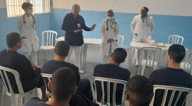 Foto mostra uma sala de aula do centro, com paredes nas cores branca e azul. Nela há cinco adolescentes sentados em cadeiras brancas, de costas, tendo à sua frente quatro adultos vestidos com roupas de profissionais de saúde, sendo que dois deles estão com estetoscópios pendurados no pescoço. Uma mulher, a professora, gesticula enquanto explica