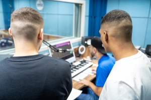 Foto mostra o estúdio de gravação na Fábrica de Cultura. Dois jovens, um branco e um negro, de pé e de costas, observam o técnico, um homem branco, sentado mexer no computador e na mesa de mixagem. Ao fundo aparece o vidro que separa a cabine de gravação da mesa técnica.