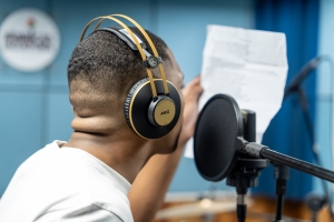 Foto mostra a cabine de gravação do estudo da Fábrica da Cultura, com o close de um jovem negro, com um fone de ouvido e cantando de frente para um microfone profissional com filtro, segurando uma folha de papel