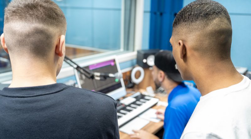 Foto mostra o estúdio de gravação na Fábrica de Cultura. Dois jovens, um branco e um negro, de pé e de costas, observam o técnico, um homem branco, sentado mexer no computador e na mesa de mixagem. Ao fundo aparece o vidro que separa a cabine de gravação da mesa técnica.