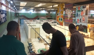 Foto mostra três jovens no piso superior, observando a parte inferior do prédio. É um grande saguão, com uma estrutura arquitetônica com paredes brancas brancas e portas