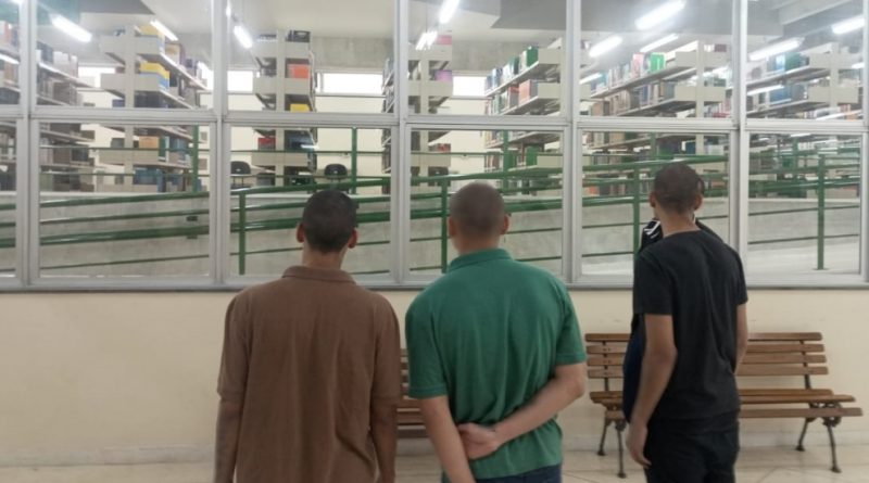 Foto mostra três adolescentes, acompanhados de um adulto, observando do lado de fora a biblioteca do Instituto. Através das janelas de vidro é possível ver prateleiras com livros organizados