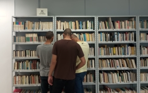 Foto mostra três adolescentes de costas, de pé à frente de prateleiras com livros organizados. dois buscam livros e um terceiro lê um deles folheado