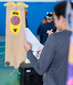 Foto mostra o close de um jovem em pé, segurando uma folha de papel com a mão esquerda, um microfone com a mão direita, e lendo o conteúdo escrito. Ele está de costas na quadra do centro socioeducativo, decorada com motivos dos livros