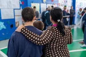 Foto mostra ao fundo um grupo de mais de cinco jovens sobre um palco, dispostos um ao lado do outro, de frente para uma parede decorada com cartazes e motivos coloridos. De costas para quem tirou a foto está uma mãe abraçada com dois filhos: um jovem atendido na Fundação e uma criança. Eles observam a apresentação do grupo sobre o palco.