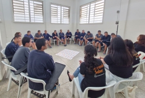 Foto mostra uma sala do centro socioeducativo, com paredes brancas. 15 adolescentes e cinco mulheres adultas estão sentados em cadeiras, em formato de roda. Ao centro, há um tecido colorido estendido no chão.