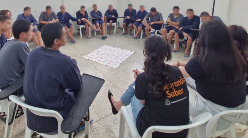 Foto mostra uma sala do centro socioeducativo, com paredes brancas. 15 adolescentes e cinco mulheres adultas estão sentados em cadeiras, em formato de roda. Ao centro, há um tecido colorido estendido no chão.