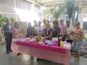 Foto mostra grupo de colaboradoras do centro socioeducativo. A frente delas, está uma mesa de café da manhã com tolhas na cor rosa