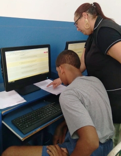 Foto mostra um jovem no laboratório de informática, sentado de frente para um computador. Ao seu lado, de pé, há uma mulher adulta, uma professora, apontando algo para o jovem em um papel escrito ao lado da tela do computador que ele utiliza