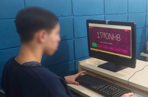 Foto mostra um adolescente no laboratório de informática do centro, sentado numa mesa, com um computador à sua frente. Na tela do computador aparece a logomarca da Olimpíada de História e o jovem utiliza o teclado.