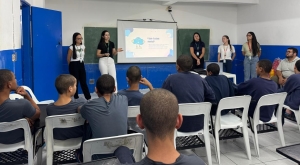Foto mostra uma sala de aula do centro de atendimento, com paredes pintadas nas cores branca e azul, tendo em uma delas uma lousa verde. Em frente à lousa há cinco mulheres de pé, palestrando para os adolescentes, havendo entre elas uma tela de projeção com slide projetado. De frente para elas e de costas para quem observa a imagem, há 9 adolescentes sentados em cadeiras, além de dois adultos