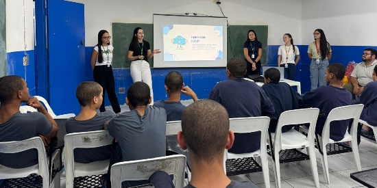 Foto mostra uma sala de aula do centro de atendimento, com paredes pintadas nas cores branca e azul, tendo em uma delas uma lousa verde. Em frente à lousa há cinco mulheres de pé, palestrando para os adolescentes, havendo entre elas uma tela de projeção com slide projetado. De frente para elas e de costas para quem observa a imagem, há 9 adolescentes sentados em cadeiras, além de dois adultos