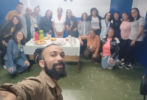 Foto mostra um grupo de 17 adultos, entre mulheres e homens, em uma das salas do centro de atendimento. Em pé, próximo à parede nas cores azul e branca, estão perfilados, como time de futebol, 16 adultos, tendo uma mesa com bolos e salgados à sua frente. Em primeiro plano aparece um homem branco, com barba, segurando uma câmera e fazendo uma selfie de todos