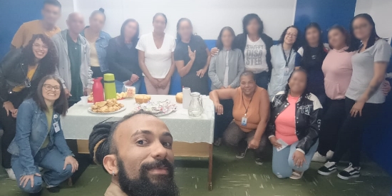 Foto mostra um grupo de 17 adultos, entre mulheres e homens, em uma das salas do centro de atendimento. Em pé, próximo à parede nas cores azul e branca, estão perfilados, como time de futebol, 16 adultos, tendo uma mesa com bolos e salgados à sua frente. Em primeiro plano aparece um homem branco, com barba, segurando uma câmera e fazendo uma selfie de todos