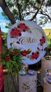 Foto retrata decoração. No centro, está um painel redondo com a palavra "Mãe" escrita em dourado, dentro de um coração com rosas vermelhas.