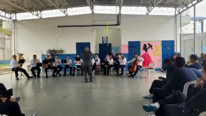 Fotografia retrata apresentação da orquestra. Os instrumentistas estão sentados em cadeiras, formando um semicírculo. Adolescentes assistem à apresentação musical