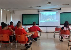 Jovens da Fundação CASA, vestidos com camisetas vermelhas, assistem a um vídeo institucional sobre a USP. Eles estão sentados em cadeiras laranjas  dentro de uma sala com paredes brancas. Ao fundo, o vídeo é reproduzido em um telão branco