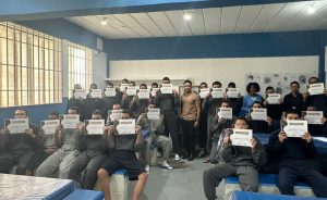 Fotografia retrata grupo de jovens que concluíram a oficina, segurando os certificados em frente ao seu rosto. No centro da foto, está o artista plástico Eduardo Carvalho 