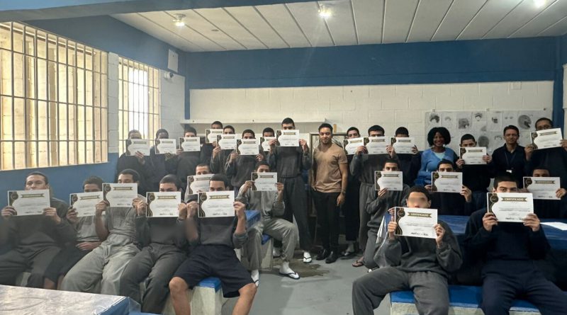 Fotografia retrata grupo de jovens que concluíram a oficina, segurando os certificados em frente ao seu rosto. No centro da foto, está o artista plástico Eduardo Carvalho