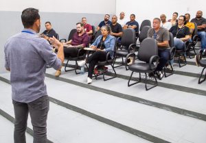 Fotografia ilustra colaboradores da Fundação CASA participando de aula em ambiente acadêmico. Profissionais de ambos os sexos, sentados em carteiras, assistem a explicação de um orientador, dentro de uma sala de aula