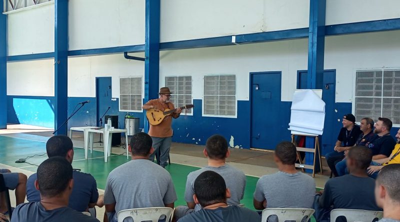 Fotografia retrata adolescentes, usando moletons e camisetas azuis, sentados em cadeiras brancas, dispostas em semicírculo. No centro, está um violeiro, que usa um chapéu marrom e barba branca. A apresentação é realizada na quadra do centro, que é coberta.