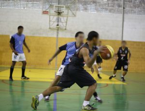 Foto retrata jovens durante partida de basquete. Em primeiro plano, dois adolescentes disputam a bola para tentar marcar uma cesta. Outros três jovens estão ao fundo da foto, fazendo a defesa da cesta. 