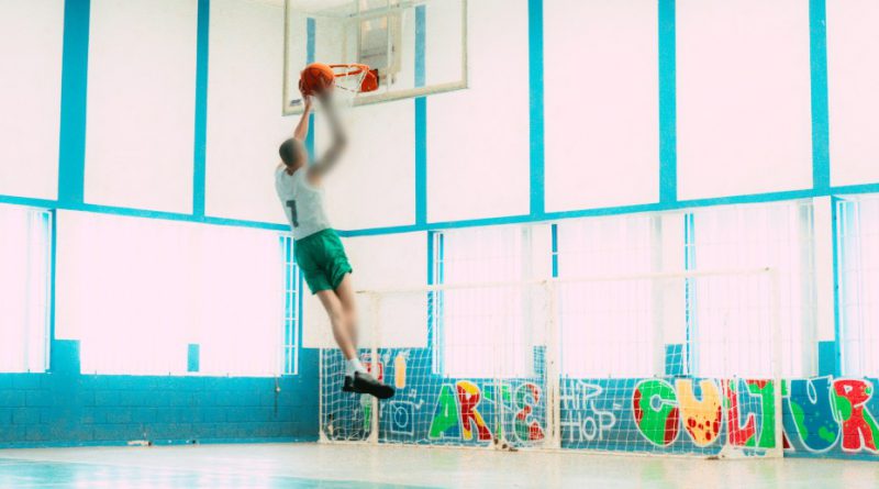 Fotografia retrata adolescente marcando uma cesta durante treino no jogo de basquete. O jovem esta próximo da tabela, após pular para marcar o ponto.