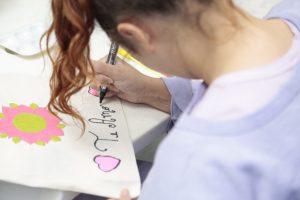 Fotografia retrata adolescente do sexo feminino, com cabelos longos, de cor acobreada e com ondas, escrevendo em uma cartolina com uma caneta do tipo marcador. Ela escreve a frase "te amo", ladeada por dois corações rosa. A jovem usa um moletom lilás.  