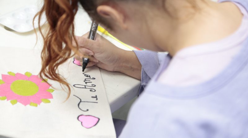 Fotografia retrata adolescente do sexo feminino, com cabelos longos, de cor acobreada e com ondas, escrevendo em uma cartolina com uma caneta do tipo marcador. Ela escreve a frase "te amo", ladeada por dois corações rosa. A jovem usa um moletom lilás.