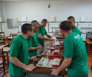 Fotografia ilustra um grupo de adolescentes, todos usando camisetas verdes, em volta de uma bancada com itens como fios, chaves tipo philips, fitas isolantes, dentre outras ferramentas utilizadas na manutenção de circuitos elétricos. 