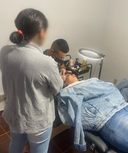 Fotografia mostra adolescente participando de uma das aulas do curso. Ela está de costas para o fotografo, usa uma blusa cinza e calça jeans. Ela observa o professor do curso realizar o procedimento em uma outra mulher, que está deitada em uma maca. 