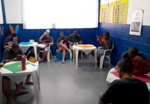 Fotografia retrata um grupo de adolescentes durante momento de leitura na biblioteca do centro. O ambiente é claro e arejado. Os jovens estão sentados em cadeiras brancas e pufs. Eles leem de forma descontraída os exemplares do livro. 