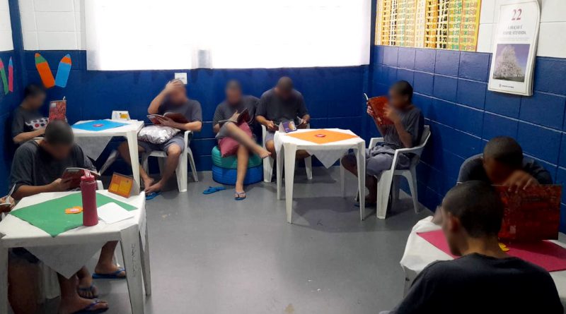 Fotografia retrata um grupo de adolescentes durante momento de leitura na biblioteca do centro. O ambiente é claro e arejado. Os jovens estão sentados em cadeiras brancas e pufs. Eles leem de forma descontraída os exemplares do livro.