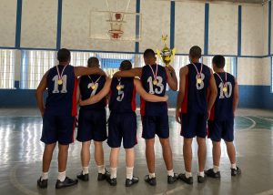 Fotografia retrata seis adolescentes, todo de costas. Eles usam uniformes em tons de azul escuro com uma listra vermelha nas laterais e se abraçam. Em suas costas, estão penduradas medalhas douradas e um deles segura o troféu de campeão. 