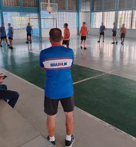 Fotografia mostra adolescentes durante jogo de basquete. Eles utilizam uniformes nas cores laranja e azul para diferenciar as equipes. Ao centro da foto, o palestrante André Brazolin acompanha a partida. Ele usa uma camiseta azul, com o nome instituto no centro, em uma faixa branca