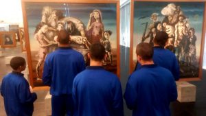 Fotografia retrata grupo de adolescentes, utilizando moletons azuis, observando a obra "Retirantes", do pintor Cândido Portinari. A obra retrata famílias de pessoas, pintadas de forma esquelética, para representar a seca que maltrata o povo nordestino. 