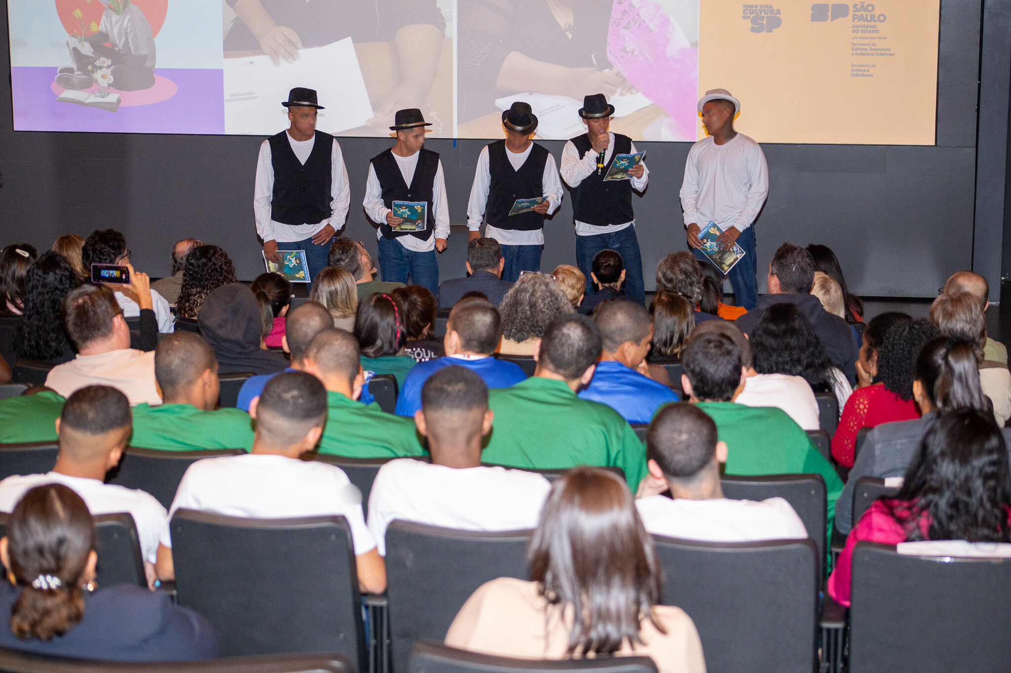 Seis adolescentes vestidos com camisas brancas e coletes pretos se apresentam no palco, segurando livros, diante de uma plateia no auditório.